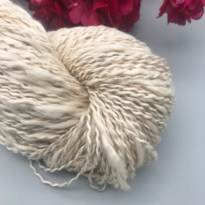 Op de afbeelding: Een close-up van een bolletje gebroken witte wol met subtiele bruine en grijze strengen. Het garen heeft een dikke, getextureerde uitstraling, wat duidt op een grof brei- of haakproject. De afbeelding is geplaatst tegen een zachte blauwe achtergrond met roze bloemen.