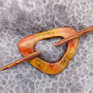 Könnte beinhalten: Eine Holzschalspange mit einem einzigartigen, abstrakten Design in Orange-, Gelb- und Brauntönen. Die Spange hat die Form einer Träne und ist glatt und poliert.