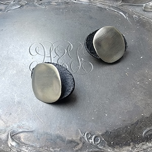 Może przedstawiać: Dwie srebrne kolczyki sztyfty w owalnym kształcie z wykończeniem ze szczotkowanego metalu. Każdy kolczyk osadzony jest na małej, okrągłej, czarnej podstawie. Kolczyki są prezentowane na srebrnej, teksturowanej powierzchni z grawerowanymi detalami.