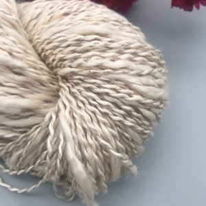 Op de afbeelding: Close-up van een bolletje gebroken witte wol met subtiele grijze en bruine vlekjes. Het garen heeft een dikke, getextureerde uitstraling, wat wijst op een chunky brei- of haakproject. De vezels zijn los gesponnen.