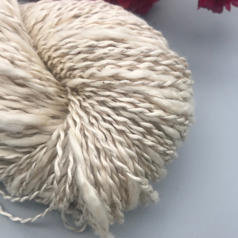 Op de afbeelding: Close-up van een bolletje gebroken witte wol met subtiele grijze en bruine vlekjes. Het garen heeft een dikke, getextureerde uitstraling, wat wijst op een chunky brei- of haakproject. De vezels zijn los gesponnen.