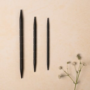 May include: Three black knitting needles of different sizes on a beige background.