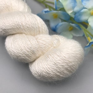 May include: Close-up of a skein of off-white yarn. The yarn is tightly wound and has a soft, textured appearance. Blue and white flowers are in the background. This yarn is suitable for knitting or crocheting projects.