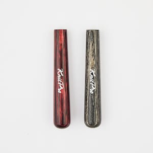 Könnte beinhalten: Zwei Stricknadel-Etuis aus Holz. Eines ist tiefrot mit Streifenmuster, das andere grau und schwarz. Der Text "Knit Pro" ist weiß auf jedem Etui aufgedruckt.