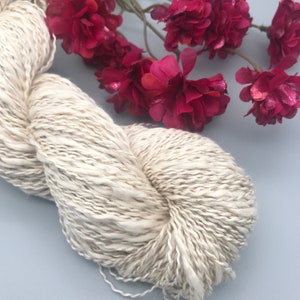 Op de afbeelding: Een streng ecru garen met subtiele grijze marmering, ideaal voor brei- of haakprojecten. Het garen is zacht en getextureerd, met een natuurlijke, organische uitstraling. Een takje dieproze bloemen voegt kleur toe aan de compositie.