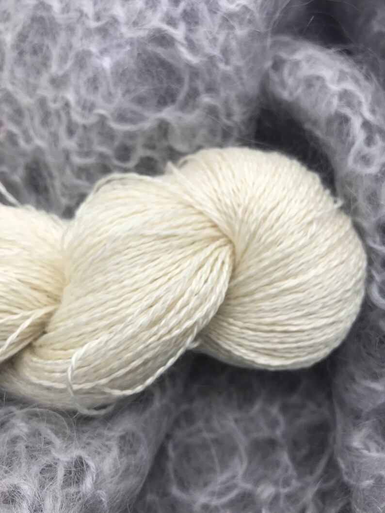 May include: A close-up of a skein of off-white yarn, tightly wound, with a soft, textured appearance. The yarn is set against a blurred background of gray, fluffy material, suggesting a knitting or crochet project.