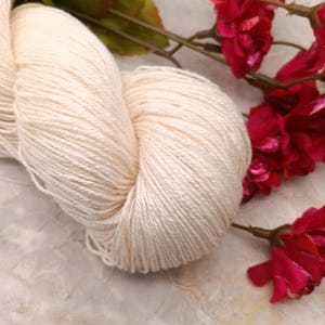 May include: A skein of off-white cotton yarn, with a soft, natural texture. The yarn is arranged in a loose coil, with the ends of the yarn visible. The yarn is photographed against a light background, with pink flowers in the background.