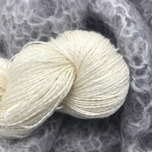 May include: A close-up shot of a skein of off-white yarn, tightly wound, with a soft, textured appearance. The yarn is set against a backdrop of fluffy, gray, and light gray yarn, creating a contrast in texture and color.