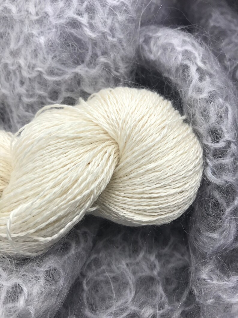May include: A close-up shot of a skein of off-white yarn, tightly wound, with a soft, textured appearance. The yarn is set against a backdrop of fluffy, gray, and light gray yarn, creating a contrast in texture and color.