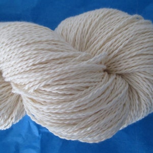 Peut inclure: Une pelote de fil blanc naturel, faite d'un m&eacute;lange de coton et de lin, est pr&eacute;sent&eacute;e sur un fond bleu.