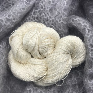 May include: Close-up of two skeins of off-white yarn, tightly wound, resting on a soft, gray, textured fabric. The yarn appears smooth and lustrous, contrasting with the fluffy background. Ideal for knitting or crochet projects.