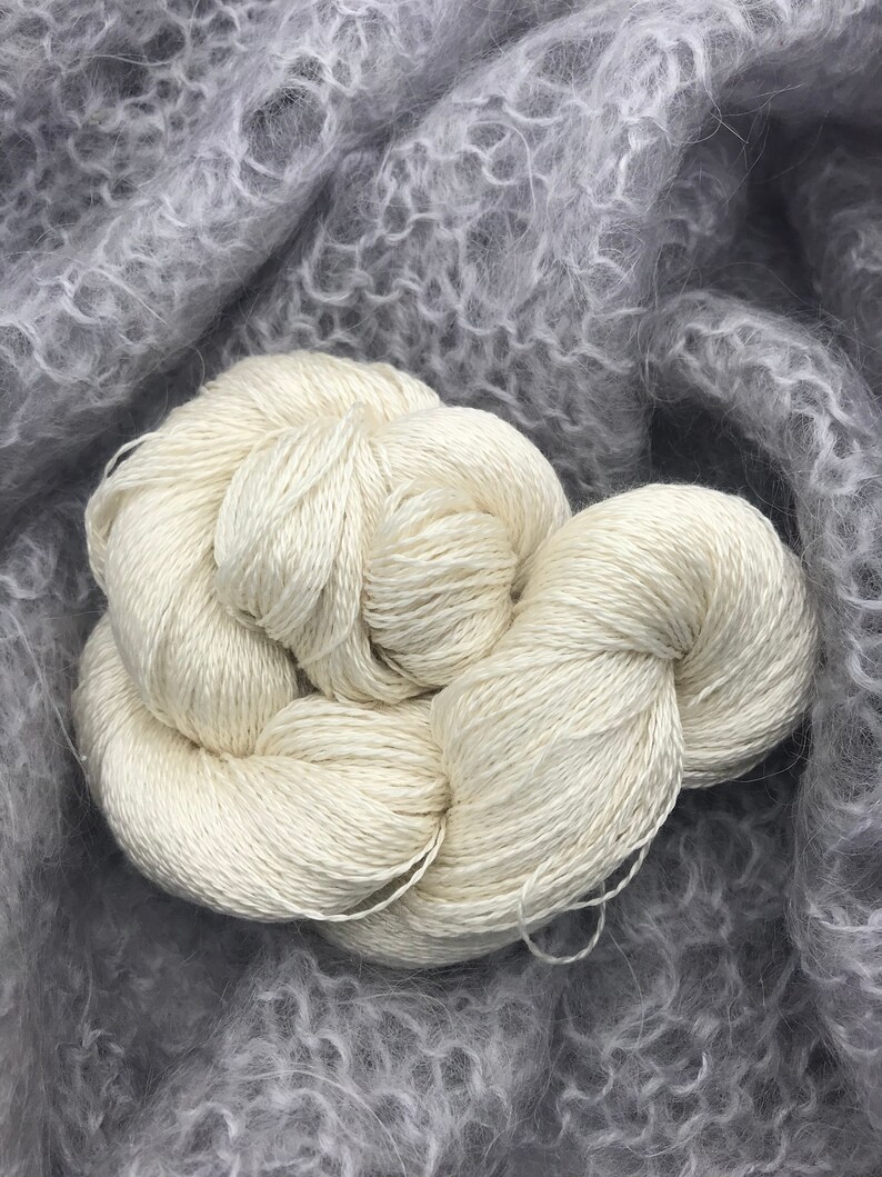 May include: Close-up of two skeins of off-white yarn, tightly wound, resting on a soft, gray, textured fabric. The yarn appears smooth and lustrous, contrasting with the fluffy background. Ideal for knitting or crochet projects.