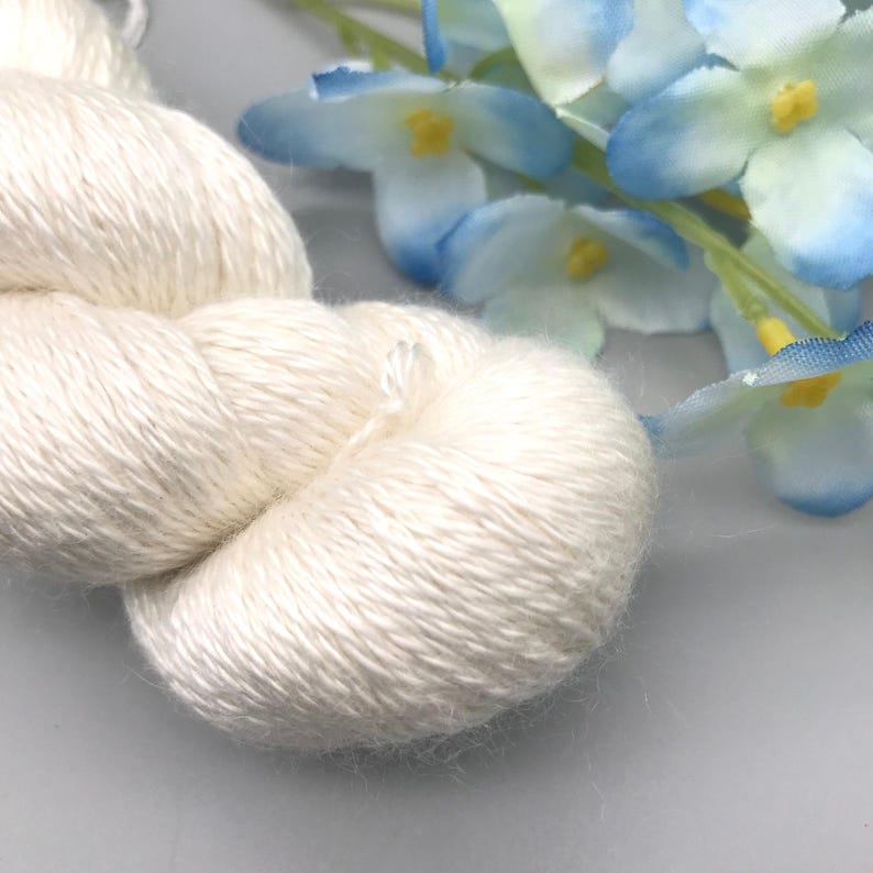 May include: Close-up of a skein of off-white yarn, tightly wound and textured. The yarn is soft and fluffy, with a slight sheen. Blue and white flowers are in the background, adding a touch of color and contrast to the neutral yarn.