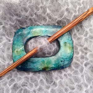 May include: A blue and green resin stitch holder with a wooden knitting needle resting inside. The stitch holder is shaped like an oval with a hole in the center. The needle is made of wood and has a natural finish.