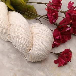 May include: A skein of white yarn, made of natural fibers, is displayed on a light gray surface. The yarn is soft and fluffy, and it is perfect for knitting or crocheting. The yarn is surrounded by red flowers.