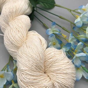 May include: A skein of natural white yarn, made from a blend of cotton and linen, is displayed against a background of blue and green artificial flowers.
