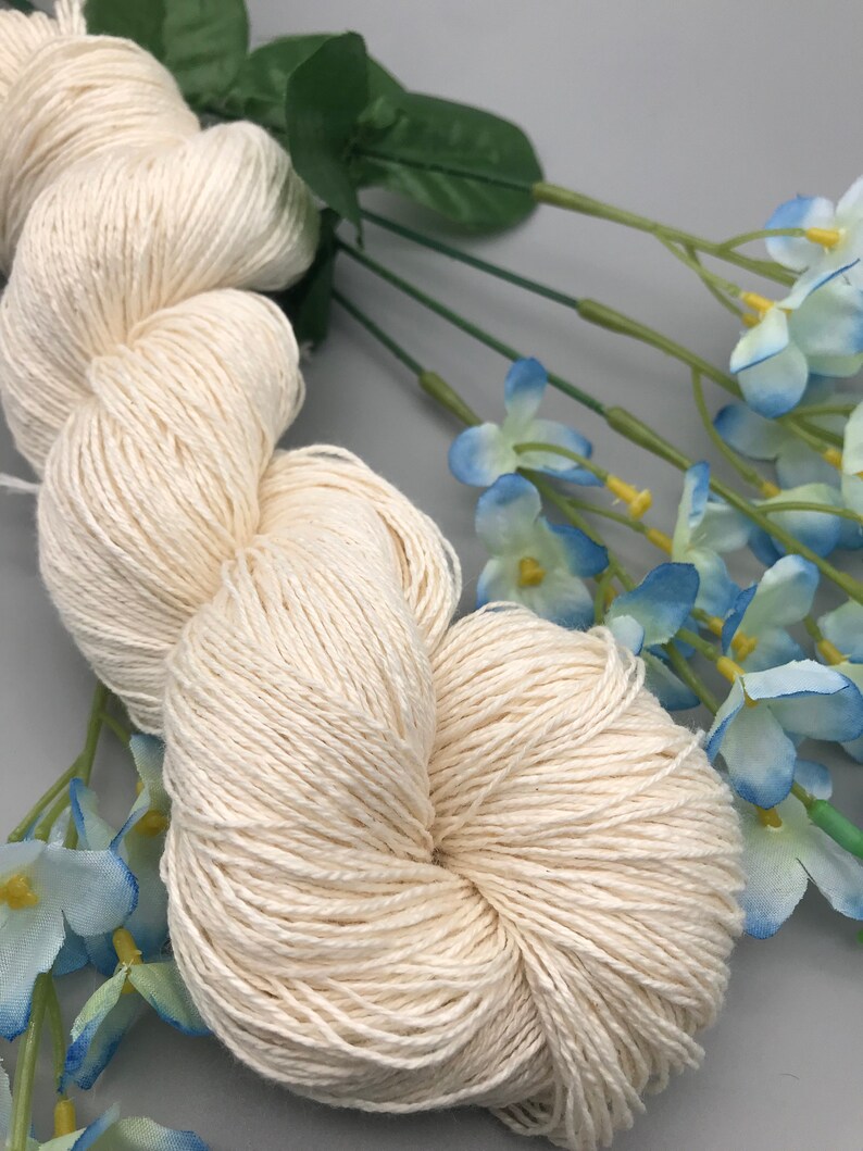 May include: A skein of natural white yarn, made from a blend of cotton and linen, is displayed against a background of blue and green artificial flowers.