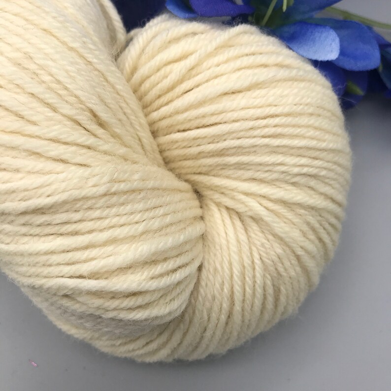 May include: A close-up of a skein of off-white yarn. The yarn is tightly wound, showing individual strands. The background includes blue artificial flowers. This yarn is suitable for knitting and crocheting projects.