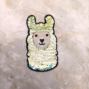 May include: A llama-shaped patch with a black outline, adorned with iridescent sequins. The llama has a cream-colored face with a heart-shaped nose and small black eyes. The sequins create a shimmering effect, adding a touch of sparkle.