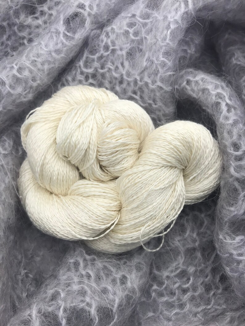 May include: A close-up shot of two skeins of off-white yarn, tightly wound and textured, resting on a soft, gray, fuzzy fabric. The yarn appears to be made of a natural fiber, possibly silk or a similar material, ideal for knitting or crochet projects.