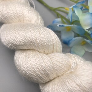 May include: A close-up of a skein of off-white yarn, tightly wound and textured. The yarn appears soft and lustrous, with a slight sheen. Blue and green artificial flowers are in the background, adding a touch of color.