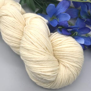 May include: A skein of off-white yarn, tightly wound, with visible texture. The yarn is a creamy color, and is partially framed by blue flowers and green leaves.
