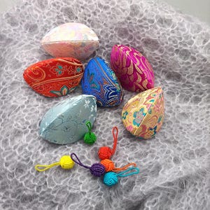 Könnte beinhalten: Eine Sammlung farbenfroher, gemusterter Stoffobjekte in verschiedenen Formen, darunter Ovale und Tropfen. Die Objekte sind in Rot-, Blau-, Rosa-, Gold- und Grüntönen gehalten und weisen aufwendige Blumen- und geometrische Designs auf. Kleine, dekorative geknotete Quasten in mehreren Farben sind ebenfalls vorhanden.
