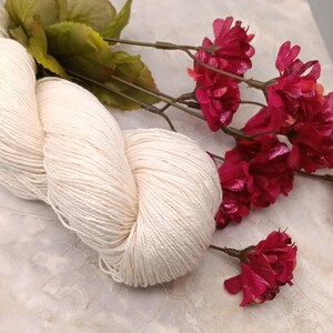 May include: A skein of natural white yarn, with a soft, fluffy texture, is displayed on a light gray surface. A bouquet of bright pink flowers with metallic accents is arranged next to the yarn.