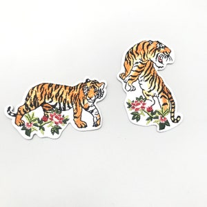 Può includere: Due toppe ricamate da stirare con tigri a strisce arancioni e nere, circondate da foglie verdi e fiori rossi.