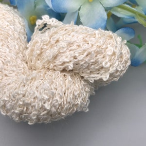 May include: A close-up of a skein of white yarn with a fluffy, textured appearance. The yarn is tightly wound and appears to be made of a soft, delicate material.