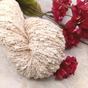 Puede incluir: Un ovillo de hilo texturizado blanquecino con una superficie en bucle y nudosa. El hilo está colocado junto a una ramita de flores rojas con tallos verdes, creando un contraste de texturas y colores. El hilo parece suave y acogedor.