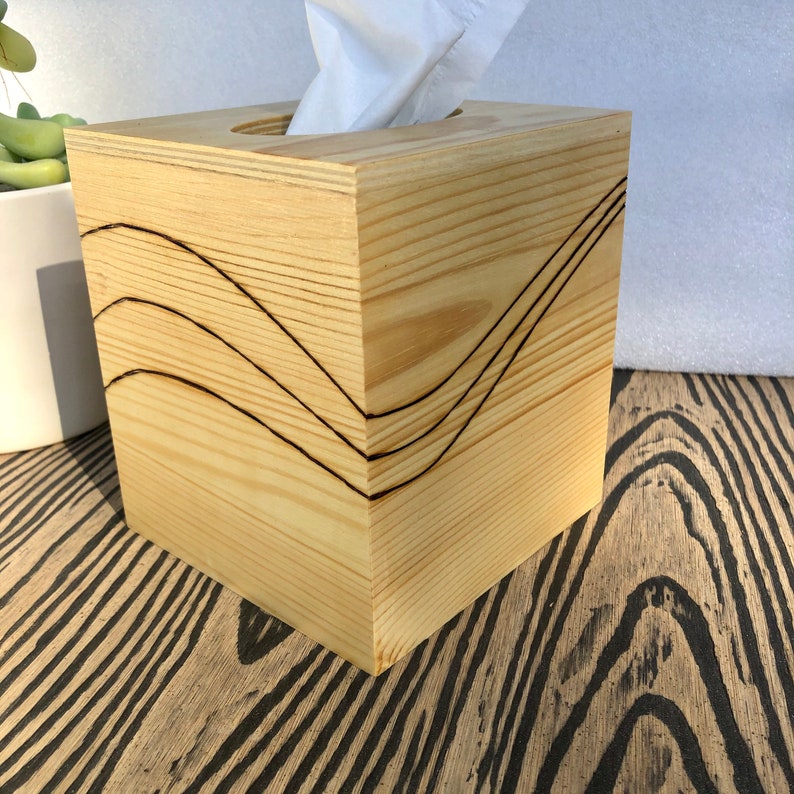 Pode incluir: Uma capa de caixa de len&ccedil;os de papel em madeira com acabamento em madeira natural e um design decorativo queimado em madeira. O design apresenta tr&ecirc;s linhas curvas.
