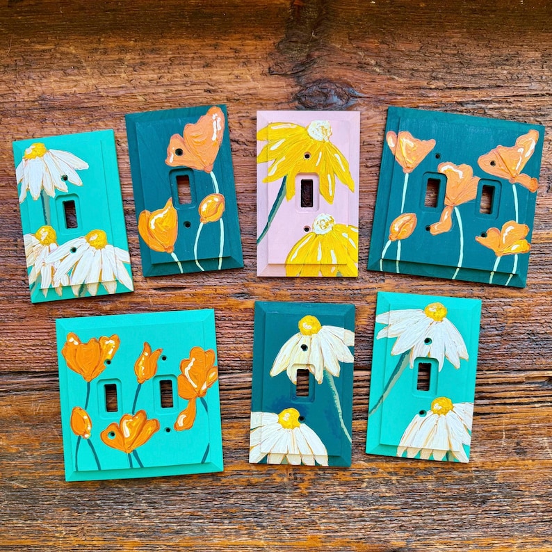 K&ouml;nnte beinhalten: Eine Sammlung von sieben rechteckigen Lichtschalterabdeckungen mit handgemalten Blumenmotiven. Die Abdeckungen sind in Blaugr&uuml;n, Hellrosa und Wei&szlig; gehalten und mit orangefarbenen und wei&szlig;en Blumenmustern verziert. Jede Platte hat Ausschnitte.