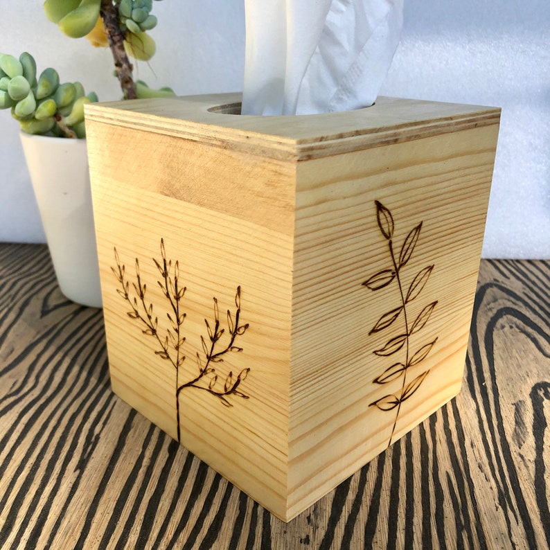 Pode incluir: Uma capa de caixa de len&ccedil;os de papel em madeira com acabamento em madeira natural. A caixa tem formato quadrado e apresenta dois desenhos simples de folhas gravadas a laser na frente e no lado.