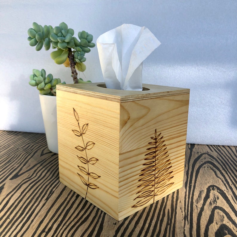 Pode incluir: Um porta-len&ccedil;os de madeira com acabamento em madeira natural. A caixa tem um recorte para os len&ccedil;os e apresenta um design queimado de dois ramos frondosos na frente.