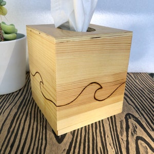 Pode incluir: Uma tampa de caixa de len&ccedil;os de papel em madeira com um design de onda queimado na madeira. A caixa &eacute; feita de madeira clara e tem um acabamento natural.