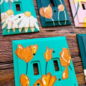 K&ouml;nnte beinhalten: Eine Sammlung rechteckiger Lichtschalterabdeckungen mit handgemalten Blumenmotiven. Die Abdeckungen sind in T&uuml;rkis, Dunkelgr&uuml;n und Hellrosa gehalten und zeigen wei&szlig;e G&auml;nsebl&uuml;mchen sowie orangefarbene Mohnblumen mit sichtbarem Pinselstrich.