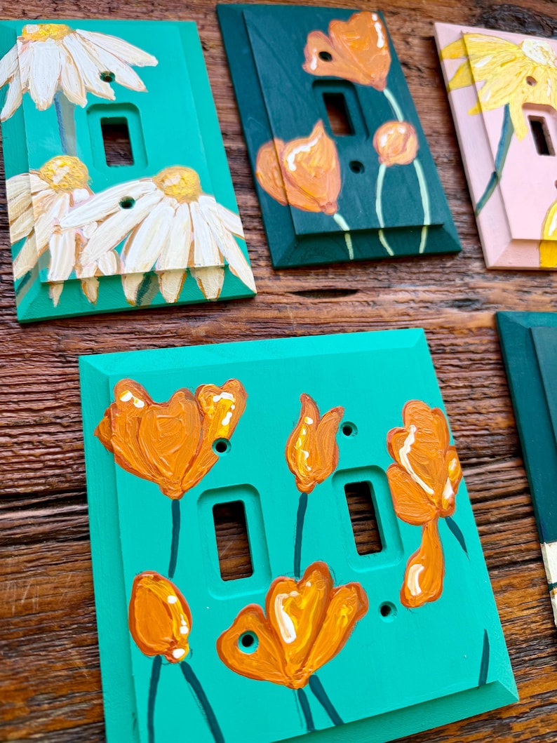 K&ouml;nnte beinhalten: Eine Sammlung rechteckiger Lichtschalterabdeckungen mit handgemalten Blumenmotiven. Die Abdeckungen sind in T&uuml;rkis, Dunkelgr&uuml;n und Hellrosa gehalten und zeigen wei&szlig;e G&auml;nsebl&uuml;mchen sowie orangefarbene Mohnblumen mit sichtbarem Pinselstrich.