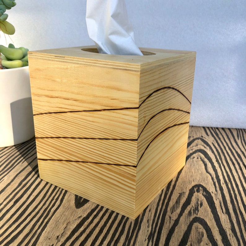 Pode incluir: Uma caixa de len&ccedil;os de papel de madeira com acabamento em madeira natural e um design decorativo queimado em madeira. A caixa &eacute; quadrada e tem uma abertura retangular para os len&ccedil;os.