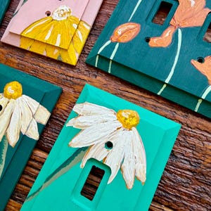 K&ouml;nnte beinhalten: Eine Sammlung rechteckiger Lichtschalterabdeckungen mit handgemalten Blumenmotiven. Die Abdeckungen sind in verschiedenen Farben wie Rosa, Dunkelgr&uuml;n und leuchtendem T&uuml;rkis gehalten und mit strukturierten gelben, orangen oder wei&szlig;en Blumen verziert.