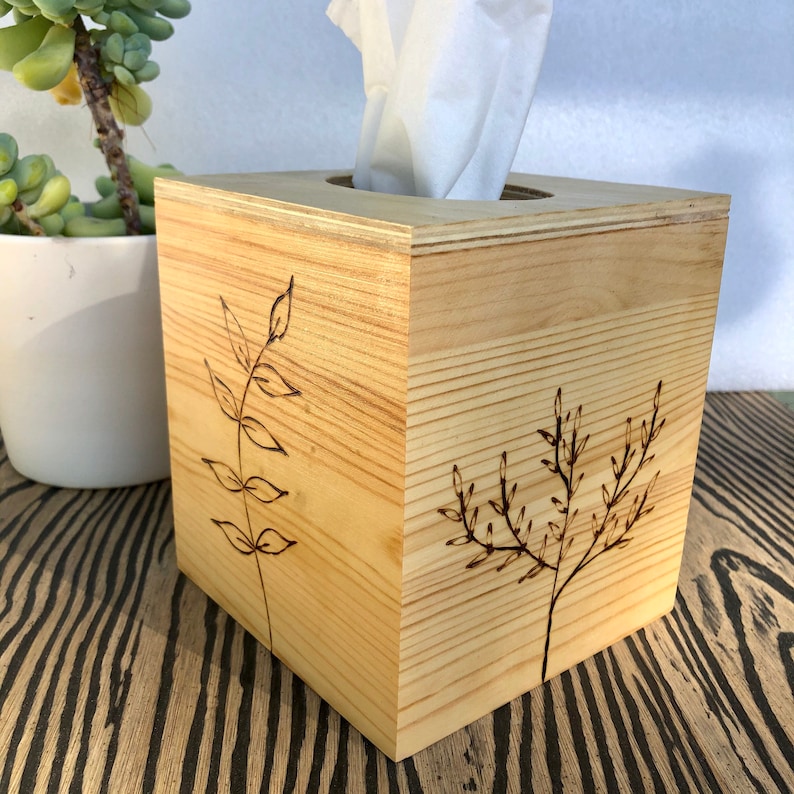 Pode incluir: Uma capa de caixa de len&ccedil;os de papel em madeira com acabamento em madeira natural. A caixa tem formato quadrado e apresenta um design simples de dois ramos com folhas queimadas na madeira.