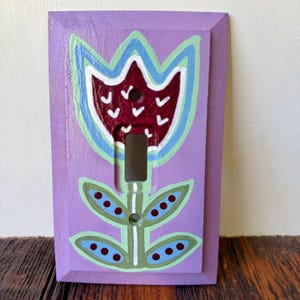 K&ouml;nnte beinhalten: Eine rechteckige Lichtschalterabdeckung in Lavendel mit einem handgemalten Blumenmotiv. Die Blume hat eine dunkelrote Mitte mit wei&szlig;en V-f&ouml;rmigen Markierungen, umrandet in Hellblau und Gr&uuml;n, mit gr&uuml;nen Bl&auml;ttern und dunkelroten Punkten.