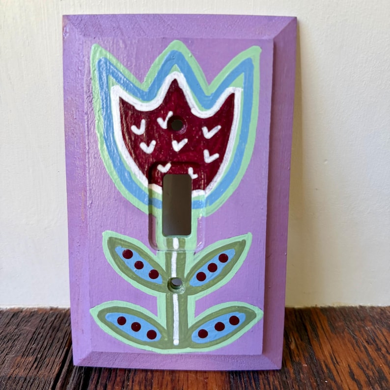 K&ouml;nnte beinhalten: Eine rechteckige Lichtschalterabdeckung in Lavendel mit einem handgemalten Blumenmotiv. Die Blume hat eine dunkelrote Mitte mit wei&szlig;en V-f&ouml;rmigen Markierungen, umrandet in Hellblau und Gr&uuml;n, mit gr&uuml;nen Bl&auml;ttern und dunkelroten Punkten.