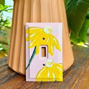 K&ouml;nnte beinhalten: Eine rechteckige Lichtschalterabdeckung mit hellrosa Hintergrund und zwei gelben Blumen mit wei&szlig;en Zentren. Das Design verf&uuml;gt &uuml;ber eine zentrale vertikale &Ouml;ffnung f&uuml;r einen Kippschalter sowie zwei Schraubenl&ouml;cher, eines oberhalb und eines unterhalb.