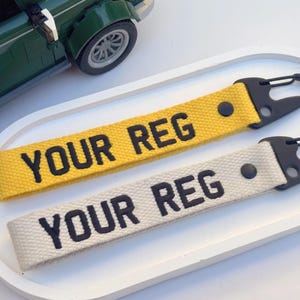 Puede incluir: Dos llaveros con el texto "YOUR REG" en negro. Uno es amarillo, el otro blanquecino. Ambos tienen cierres de metal negro. Un coche de juguete verde está en el fondo.