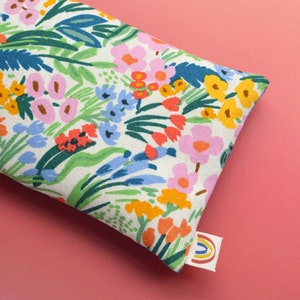 May include: A white fabric eye pillow with a colorful floral print. The pillow has a rainbow-colored label attached to it.