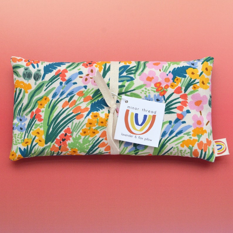 May include: A white fabric lavender and flax pillow with a floral print in shades of yellow, orange, pink, blue, and green. The pillow is tied with a white ribbon and has a tag that reads "minor thread lavender & flax pillow".