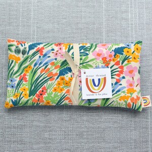 May include: A white fabric lavender and flax pillow with a floral print in shades of pink, orange, yellow, blue, and green. The pillow is tied with a cream ribbon and has a tag that reads "minor thread" with a rainbow logo and the text "lavender & flax pillow".