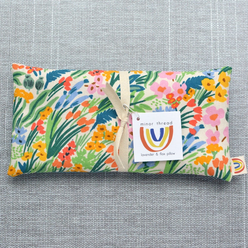 May include: A white fabric lavender and flax pillow with a floral print in shades of pink, orange, yellow, blue, and green. The pillow is tied with a cream ribbon and has a tag that reads "minor thread" with a rainbow logo and the text "lavender & flax pillow".