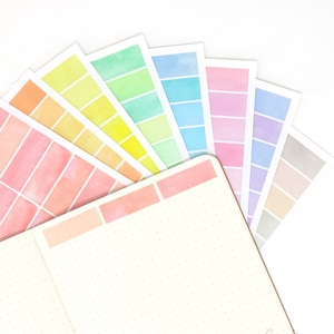 May include: A set of pastel coloured watercolour planner stickers with a grid pattern. The stickers are arranged in a fan shape and are on a white background.